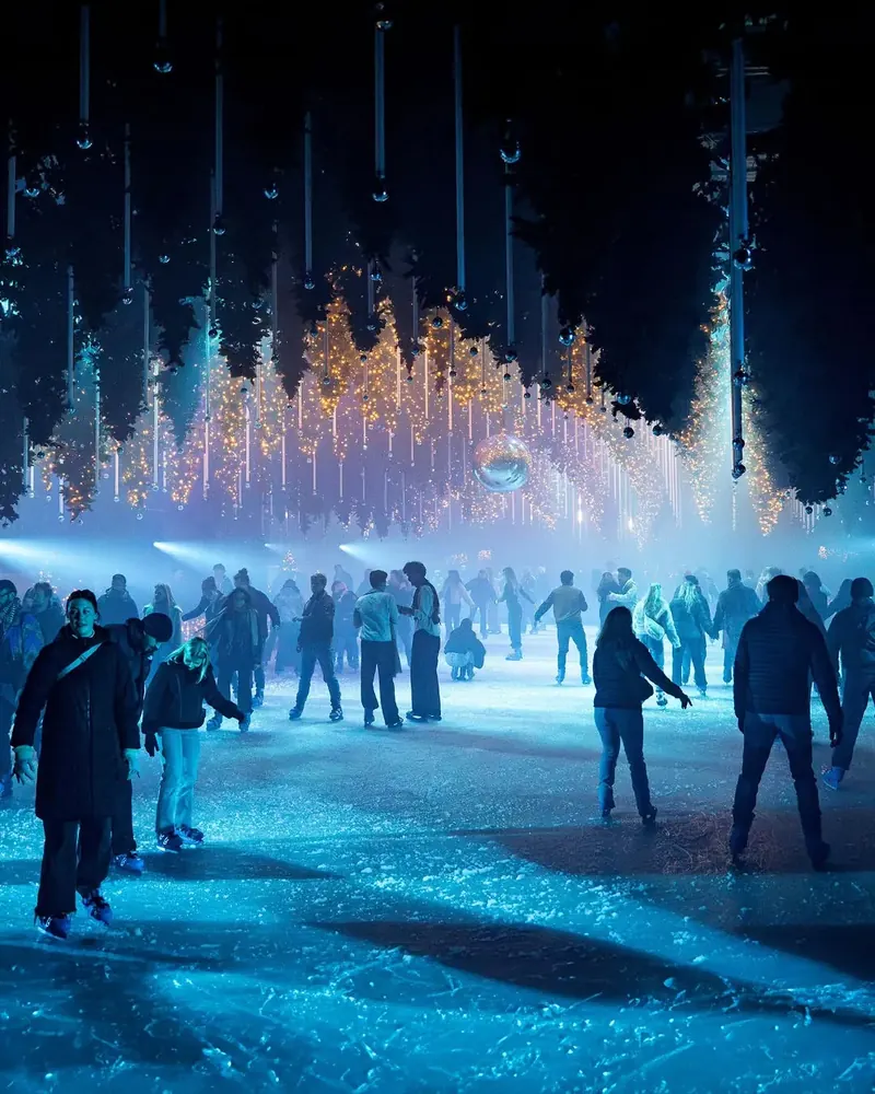 Skaters on an ice rink lit blue beneath hanging trees with Christmas light strings