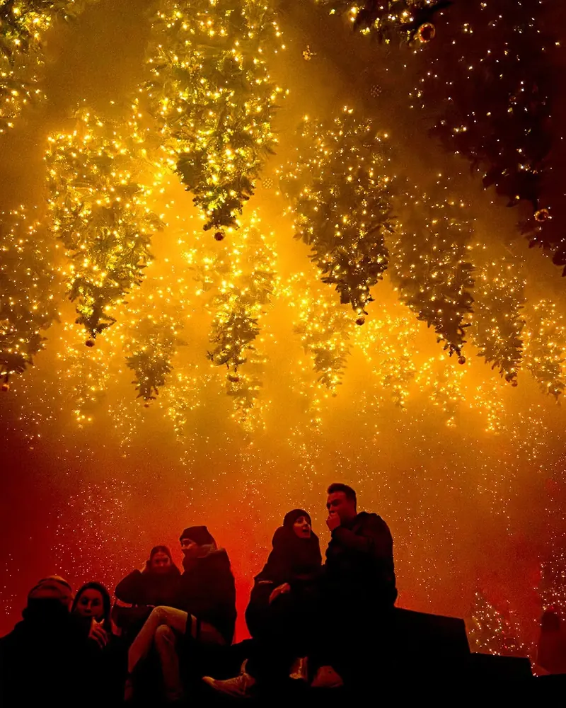 Visitors resting under hanging Christmas trees with string lights in a golden haze